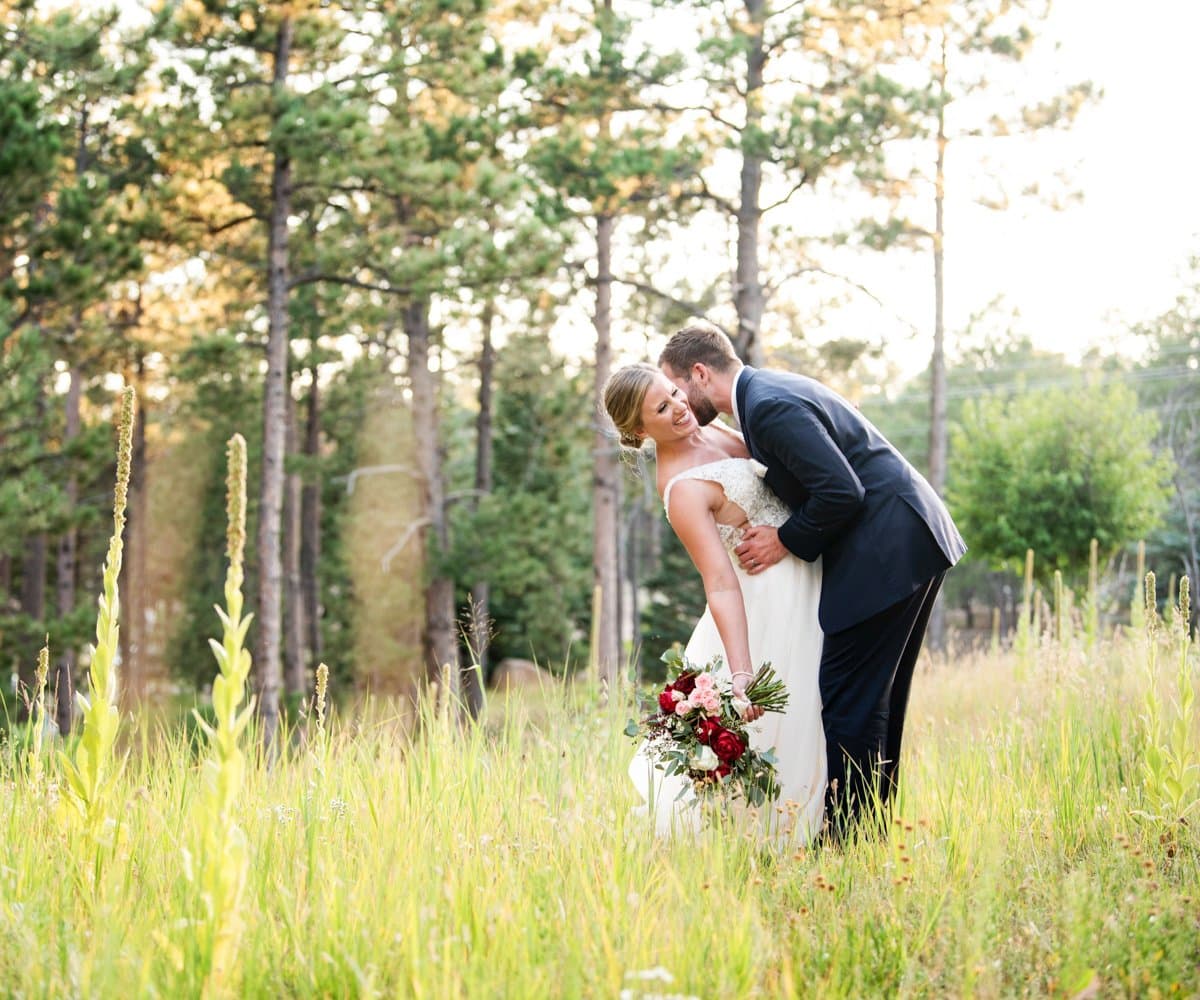 Bride and groom sharing a kiss in a sunlit meadow framed by pines at Wedgewood Black Forest in Black Forest, Colorado