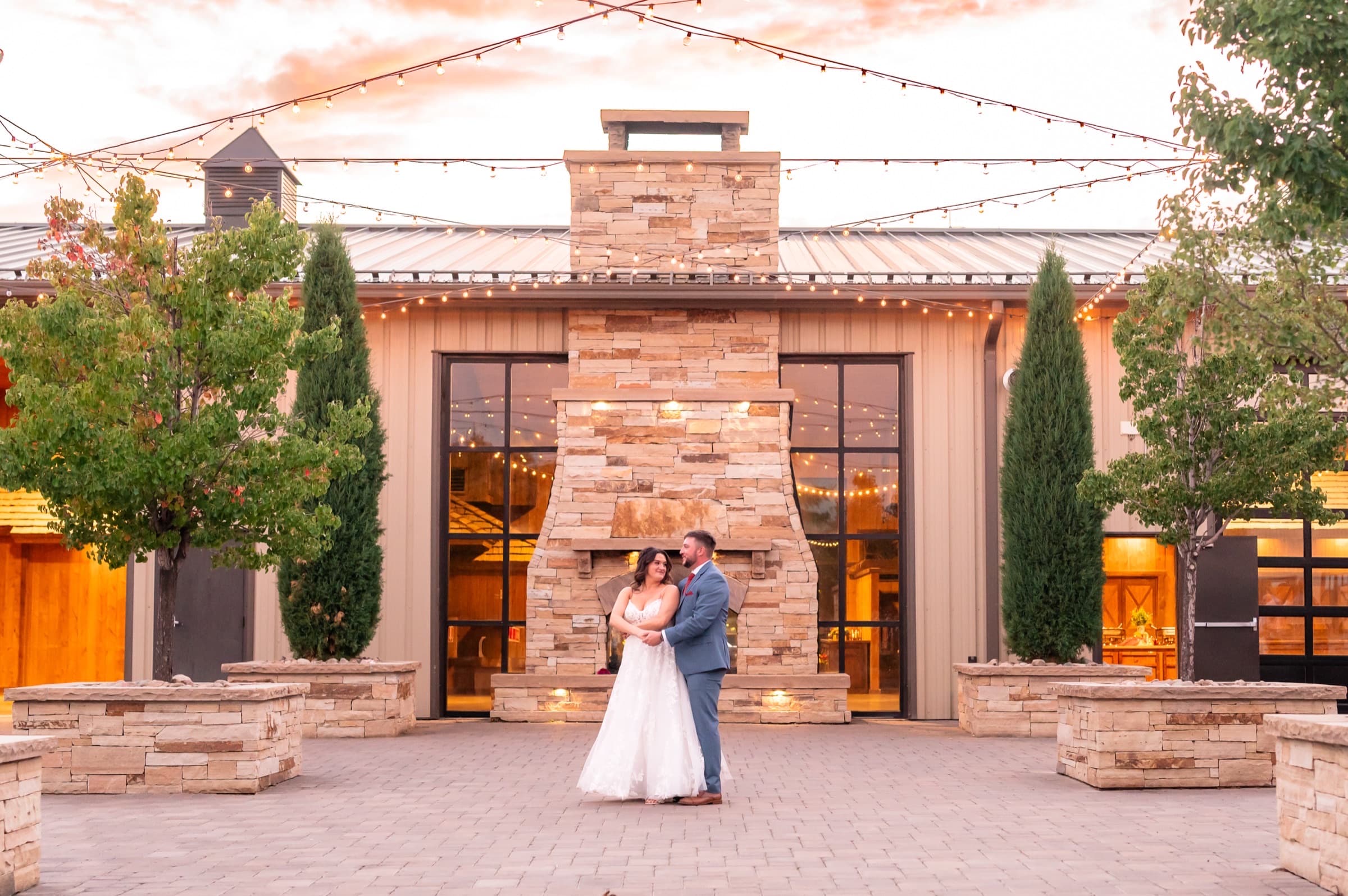 Bride and groom embracing under string lights by the stone fireplace at sunset at Spruce Mountain Ranch in Larkspur, Colorado